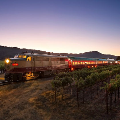 A vintage train labeled "Napa Valley" travels through a vineyard at dusk, with a mountain silhouette in the background.
