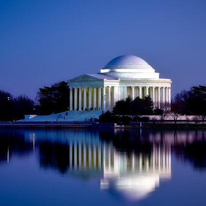 The Jefferson Memorial illuminated at night, reflected in a calm body of water, with trees and a clear evening sky.