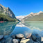 A tranquil lake with mountains and trees reflected in the clear water, rocks in the foreground, and a dock with boats on the left.