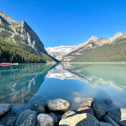 A tranquil lake with mountains and trees reflected in the clear water, rocks in the foreground, and a dock with boats on the left.
