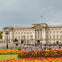 A large historic building with a flag on top, a fountain, and colorful flowerbeds in the foreground under a cloudy sky.