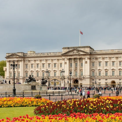A large historic building with a flag on top, a fountain, and colorful flowerbeds in the foreground under a cloudy sky.