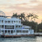 A modern white building with large windows and a dome roof sits by the water, surrounded by palm trees during sunset.