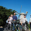 Four people with bicycles standing in front of a windmill, two of them wearing helmets. Trees and blue sky in the background.