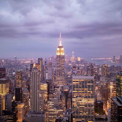 A cityscape view of Manhattan at dusk, featuring the Empire State Building illuminated against a cloudy sky.