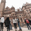 People standing and talking in front of a large, historic building with detailed architecture and a clock tower.