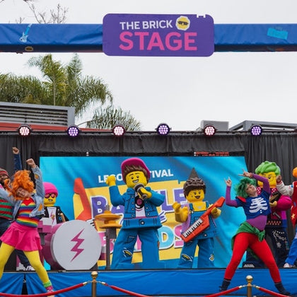 Performers in colorful costumes resembling LEGO characters dance and sing on a stage under a banner reading "The Brick Stage."