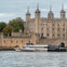 A modern boat labeled 'Uber Boat' passing by the historic Tower of London, with trees and a cloudy sky in the background.