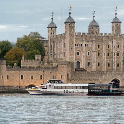A modern boat labeled 'Uber Boat' passing by the historic Tower of London, with trees and a cloudy sky in the background.