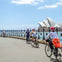 Cyclists riding along a waterfront path with the Sydney Opera House visible in the background under a clear sky.