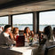 A group of people dining at a table by large windows on a boat, overlooking a scenic lake with sailboats and trees.
