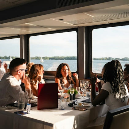 A group of people dining at a table by large windows on a boat, overlooking a scenic lake with sailboats and trees.