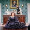 A woman in a black Victorian dress sits on the floor in front of a desk. A man in period attire stands behind the desk.