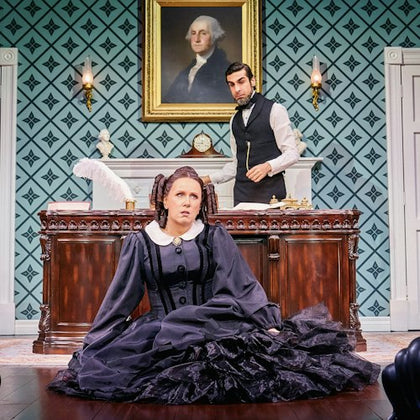 A woman in a black Victorian dress sits on the floor in front of a desk. A man in period attire stands behind the desk.