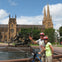 Two people wearing helmets pose with a bicycle in front of a fountain and a cathedral with twin spires on a sunny day.