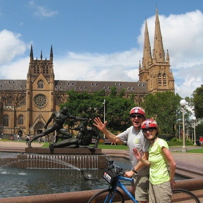 Two people wearing helmets pose with a bicycle in front of a fountain and a cathedral with twin spires on a sunny day.