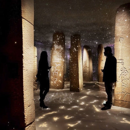Two silhouetted figures stand in a dimly lit room with tall ancient stone pillars covered in carvings and glowing lights.