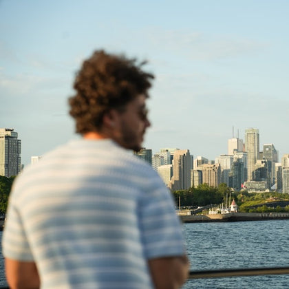 A person with curly hair and a striped shirt stands by a railing, overlooking a body of water and a city skyline at dusk.