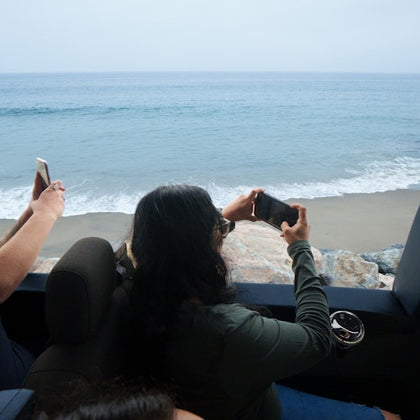 People in a vehicle by the ocean, taking photos of the beach and waves through the open window.