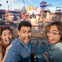 Two excited people, a woman and a man, pose with wide smiles in front of an outdoor action set with explosions in the background.