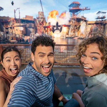 Two excited people, a woman and a man, pose with wide smiles in front of an outdoor action set with explosions in the background.