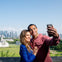 A couple smiles while taking a selfie with a city skyline and green park in the background.