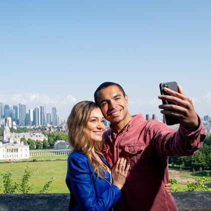 A couple smiles while taking a selfie with a city skyline and green park in the background.