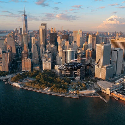 Aerial view of a helicopter flying over the waterfront near skyscrapers in downtown Manhattan at sunset.