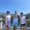 Three people in sporty attire standing on a bridge with a lush, green landscape and clear blue sky in the background.