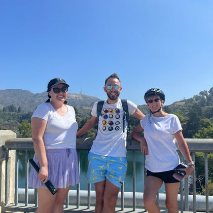Three people in sporty attire standing on a bridge with a lush, green landscape and clear blue sky in the background.