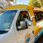 A man in a white van with yellow accents, smiling and making a peace sign, parked in a sunny urban area.