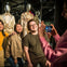 Three people posing and smiling for a photo in front of a display of mannequins wearing elaborate costumes, with another person taking their picture using a smartphone.