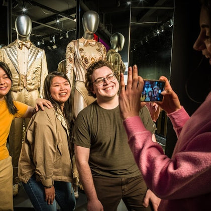 Three people posing and smiling for a photo in front of a display of mannequins wearing elaborate costumes, with another person taking their picture using a smartphone.