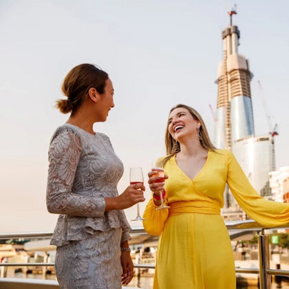 Two women in elegant dresses holding glasses and smiling on a balcony, with a city skyline and tall building under construction.