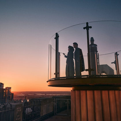 Two people stand on a circular glass observation deck at sunset, overlooking a city skyline with a clear sky.