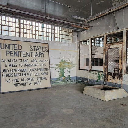 An interior view of a room with a sign reading "United States Penitentiary" and weathered walls and barred windows.
