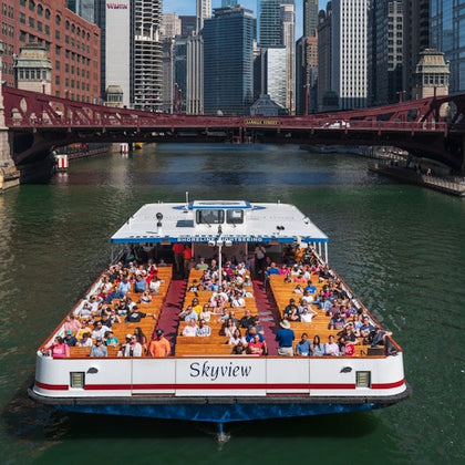 A sightseeing boat named 'Skyview' filled with people cruising on a river through a city with tall buildings and a bridge.