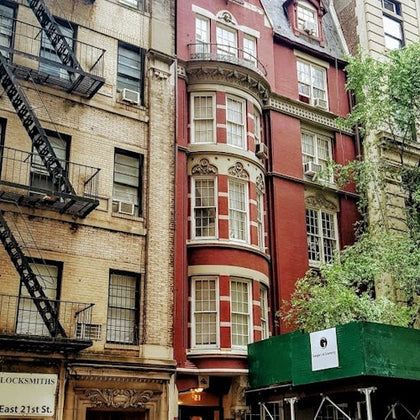 A red brick building with ornate details and curved windows, flanked by beige buildings, with a green awning at the entrance.