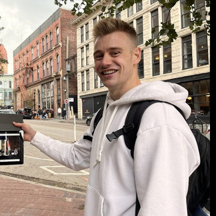 A smiling person in a white hoodie holding a tablet on a street lined with historic buildings.