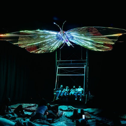 People are seated and lying on the floor under a large, glowing butterfly sculpture in a dark room.