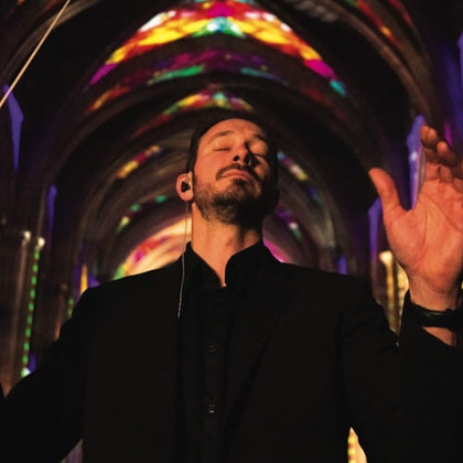 A man in a black suit conducts with closed eyes in a colorful, illuminated cathedral.