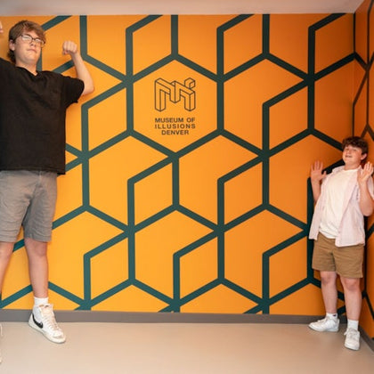 Two people posing in front of an orange wall with a geometric pattern and the text 'Museum of Illusions Denver.'
