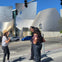 Three people stand on a street corner near a modern, metallic building with curved architectural elements under a clear blue sky.