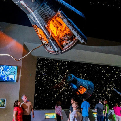 Visitors in a museum observe hanging spacecraft capsules with illuminated interiors against a starry backdrop.