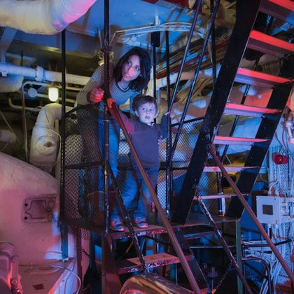 A woman and a child stand on metal stairs inside an industrial setting with pipes and machinery, lit with colored lights.