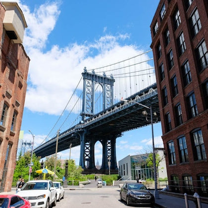 A tall blue suspension bridge viewed from a street with parked cars and brick buildings on both sides. Bright, sunny day.