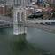A helicopter flies over the Brooklyn Bridge with city buildings and the river visible below.