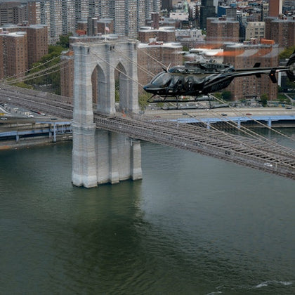 Helicopter flying near a stone tower of the Brooklyn Bridge over the East River, with city buildings in the background.