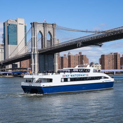 A NY Waterway ferry on the river with the Brooklyn Bridge and city buildings in the background under a clear sky.