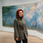 A person with red hair wearing a gray coat stands in an art gallery, looking at a large impressionist painting.